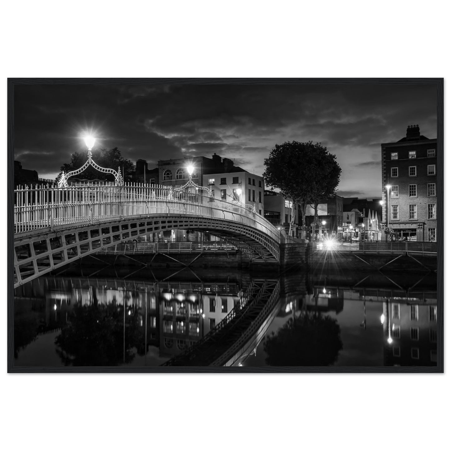 Black and white framed wall art print of the Ha'penny Bridge in Dublin at night. The bridge's historic architecture is beautifully highlighted against the dark sky, with reflections shimmering in the River Liffey below. Ideal for adding elegance to any room or office.
